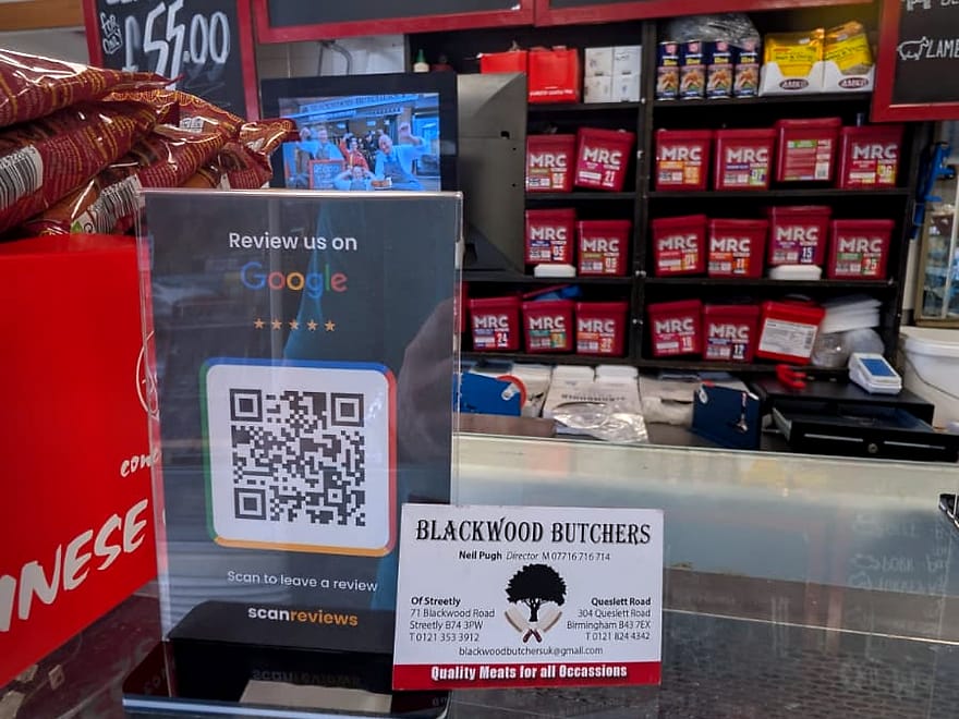 A ScanReviews Google review card standing on the counter of a butchers shop in front of the price boards